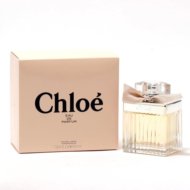 Chloe For Women Eau De Parfum 2.5 OZ with a clear ribbed glass bottle containing pale peach liquid, topped by a silver cap and beige fabric bow, displayed beside a beige box with black Chloe branding. The warm, flowery fragrance includes peonies, freesia, magnolia, rose petals, and a woody base with long-lasting freshness.