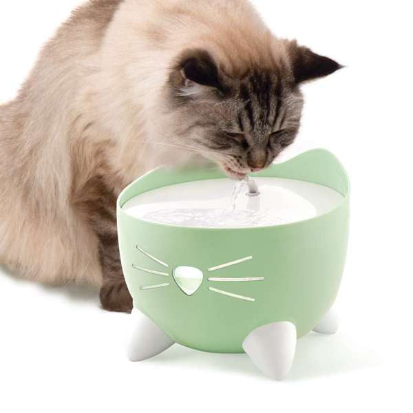 Catit PIXI Cat Fountain in green with 2.5 L capacity, featuring ergonomic cat ears and paws for easy refills, a nose-shaped backlit water level window, a quiet pump, and an arching water spout; a fluffy cat drinks from the fountain.