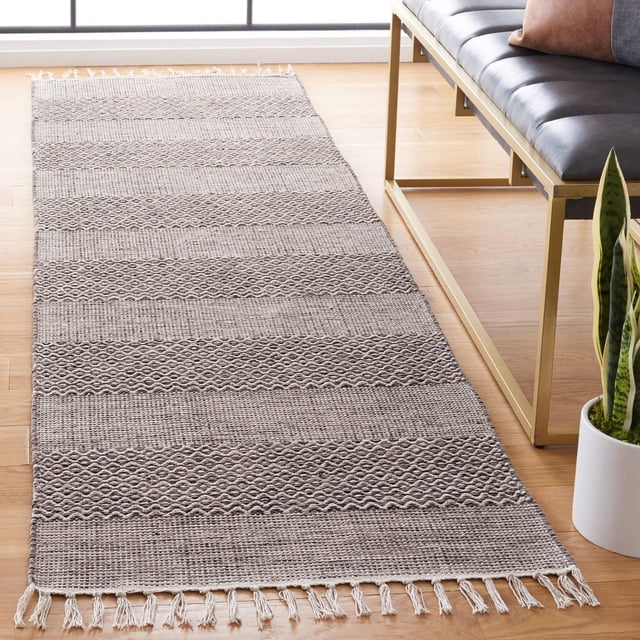 SAFAVIEH Montauk Karlene Geometric Striped Cotton Area Rug with flat-weave cotton texture, neutral beige alternating stripes and diamond patterns, rectangular runner with fringed ends on light hardwood floor beside a black leather bench and snake plant, ideal for hallways or entryways.