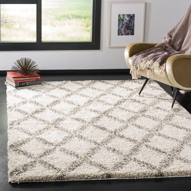 Safavieh Hudson Franklin Geometric Diamonds Shag Area Rug featuring a thick ivory shag pile with a light taupe geometric diamond pattern, set on a dark floor in a modern living room. The rug pairs with a tan armchair draped with a fringed throw and a stack of books topped by a decorative sculpture, suitable for adding soft texture to contemporary or urban décor.
