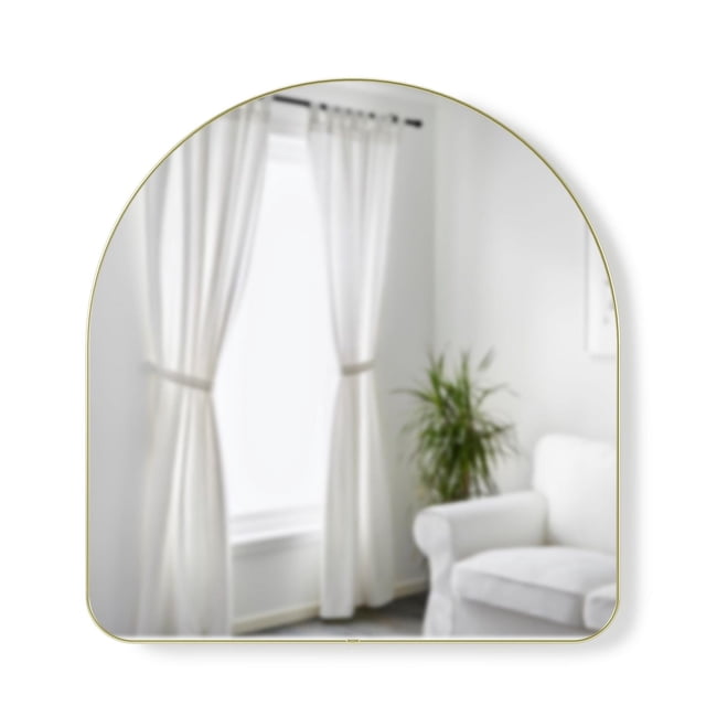 Umbra Arched 34x36" Mirror with a soft arch shape and thin brass metallic rim, reflecting a bright room with white curtains, a white armchair, and a green plant, ideal for entryway or bedroom decor and often used to enhance natural light.