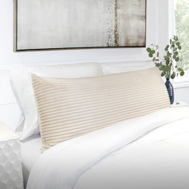 Cord Plush Body Pillow Oat with ribbed plush fabric in a soft oat colour, sized 20 by 48 inches, placed on a white bed with white pillows and bedding, complemented by a neutral abstract painting and a dark vase with eucalyptus on the bedside table, suitable for full-body support and enhancing bedroom texture.