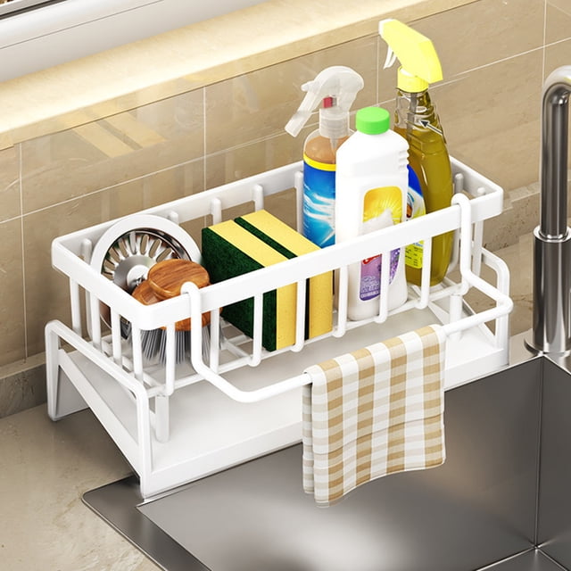 Staron Sponge Holder for Kitchen Sink, a white ABS plastic kitchen sink caddy organizer with dimensions of 9.05 inches by 4.33 inches by 3.93 inches. Designed for kitchen storage solutions, it features an inclined drainage system, towel hanging rod, and ample space for sponges, dish soap, brushes, and sink stoppers. Positioned on a kitchen counter next to a sink, holding cleaning supplies and a checkered dish towel.