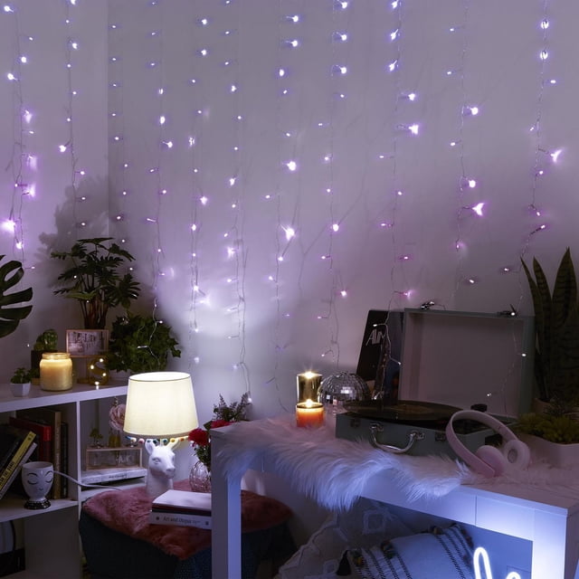Merkury Innovations XL Curtain Lights Multicolor featuring 15 strands with 240 multicolor LED lights cascading on a white wall. The flexible clear wire supports the lights above a white table draped with faux fur, holding a record player, pink headphones, and candles. Nearby, a lamp with a white shade and green plants add ambient lighting, ideal for decorative novelty lighting in home interiors.