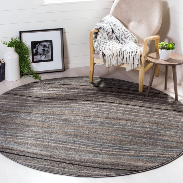 SAFAVIEH Amsterdam Aragon Striped Area Rug, round with horizontal stripes in rustic earth tones of brown, gray, and blue, made from durable synthetic yarns, shown on light hardwood floor beside a beige armchair with a white fringed throw and a small wooden side table with a potted succulent.