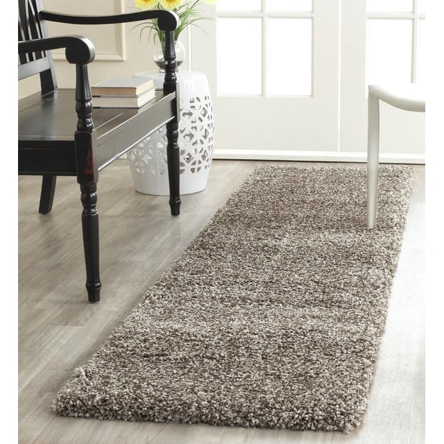 Safavieh Milan Harlow Solid Shag Area Rug in grey, a rectangular runner with dense synthetic yarns and soft shag texture, placed on light wood flooring between a black wooden chair and a white decorative side table with flowers, ideal for adding warmth and texture to contemporary home décor.