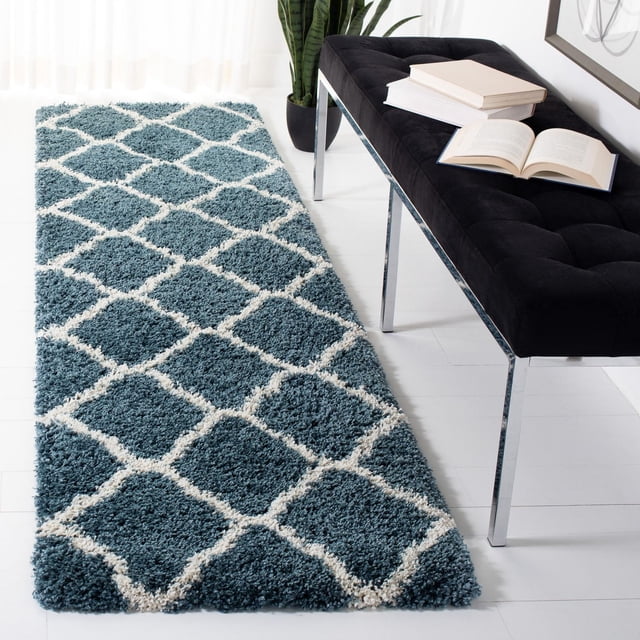 Safavieh Hudson Aline Geometric Shag Area Rug in blue with white Moroccan tile large mosaic diamond pattern, featuring a soft synthetic yarn shag pile, placed on white flooring beside a black tufted bench with chrome legs and books, in a bright room with sheer curtains and a potted plant, suitable for adding texture and warmth to runner rug spaces.