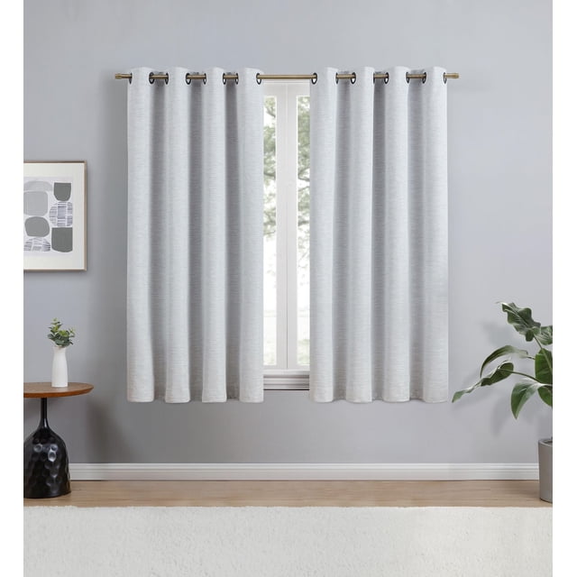Eclipse Avery 63" Grommet Blackout Window Curtains Panel in light grey hanging on a brass rod in a modern room with grey walls, wooden floor, side table with vase and plant, and a potted green plant, designed to block light, reduce noise, provide privacy, and help with energy savings.
