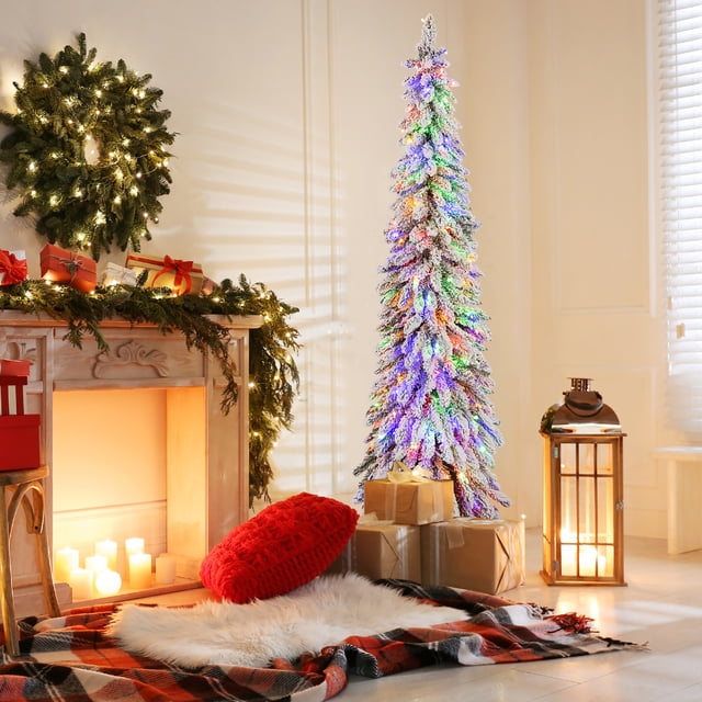Gymax 6 FT Pre-Lit Artificial Christmas Tree Snow-Flocked Slim Pencil Christmas Tree in a festive holiday room. The tree features 407 snow-flocked branch tips and 250 LED lights with warm-white and multi-color options, creating a stunning holiday centerpiece. It is surrounded by wrapped gifts, a red pillow, and cozy decor including a lit fireplace and garland.