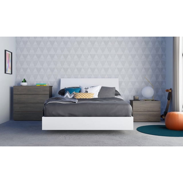 Unik 4 Piece Bedroom Set with a white floating platform bed and minimalist headboard, dressed in dark gray bedding and decorative pillows in blue, white, gray, and yellow patterns. Includes a dark wood 4-drawer chest and 2-drawer nightstand with storage drawers on metal slides. The nightstand holds a spherical lamp and digital clock. The room features a gray geometric triangle patterned wall, colorful abstract framed art, teal round rug, orange pouf, and a skateboard by the window with white curtains. This modern bedroom furniture set offers ample storage and a clean-lined profile ideal for contemporary spaces.