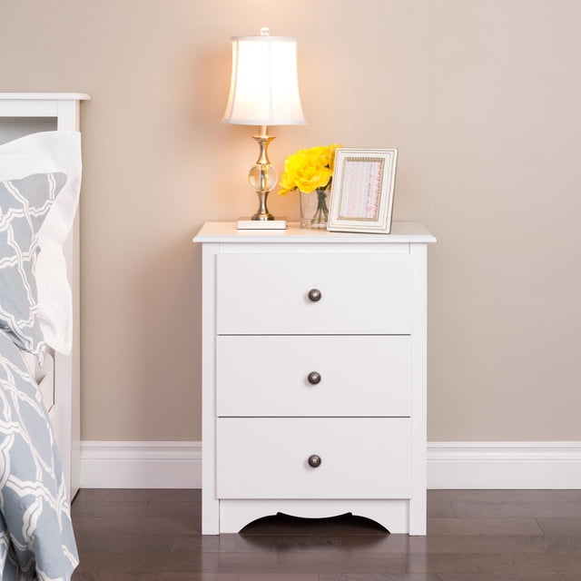Prepac table de Chevet Haute 23 Po L x 29 Po H x 16 Po D Sonoma à 3 Tiroirs, Fini Blanc, avec Trois Tiroirs Pleine Grandeur sur Glissières en Métal avec Boutons Ronds en Métal, Découpe au Bas Courbé et surface à Dessus Plat Tenant une Lampe de table en Laiton et en Verre, des Fleurs Jaunes dans un vase en Verre et une photo Encadrée, Posée sur un Plancher de Bois Franc Foncé contre un Mur beige à Côté d'Un Lit avec Literie à Motifs, Idéale pour le Rangement de Chambre et l'Organisation de Chevet.