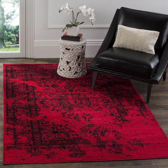 Safavieh Adirondack Zoey Traditional Faded Area Rug with red and black faded abstract traditional motifs, made of durable polypropylene yarns, placed on dark hardwood floor beside a black leather accent chair with a cream cable-knit pillow and a white ceramic garden stool holding a white orchid and books in a bright room with white paneled walls.