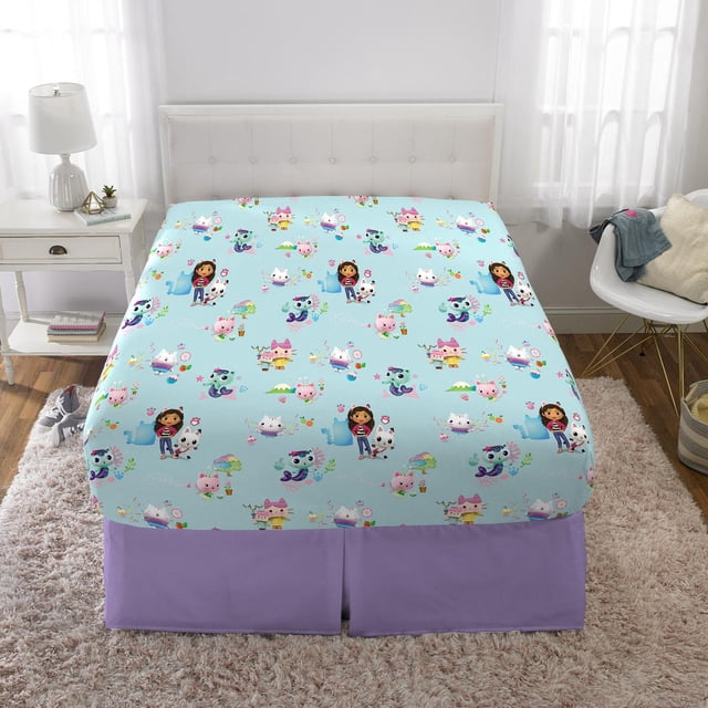 Gabby's Dollhouse "Paw Tastic" Full Sheet Set with a light blue fitted sheet featuring colorful animated characters from the DreamWorks series, including cats, a girl, and whimsical houses, on a full-size bed with a white tufted headboard in a bright bedroom with white curtains, a white nightstand with a lamp, and a purple bed skirt.