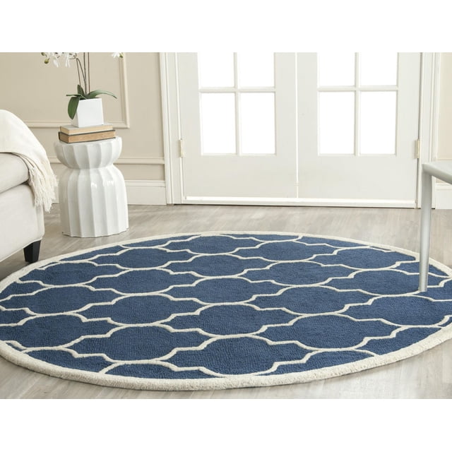 Safavieh Cambridge Kevin Geometric Area Rug, a round hand-tufted wool rug with a blue background and white geometric pattern, featuring plush cut and high/low loop pile texture, placed on light hardwood flooring near a white ceramic side table and beige sofa in a bright room, ideal for adding modern American style to home or office spaces.
