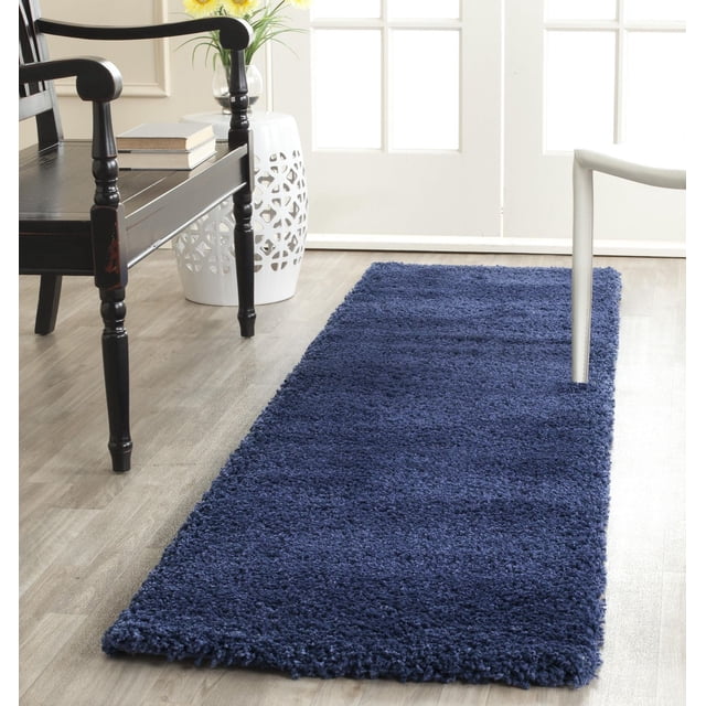 Safavieh Milan Harlow Solid Shag Area Rug in navy blue with thick shag texture, rectangular on light wood flooring between a black wooden chair and a white chair, beside a white ceramic garden stool and bright windowed door, ideal for adding warmth and texture to modern home interiors.