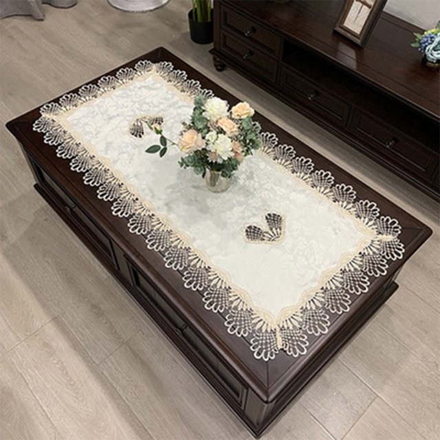 Luckyn European Embroidered Tablecloth in a rectangular shape, measuring 50x100cm, displayed on a dark wooden coffee table. The tablecloth features intricate crochet handmade lace with a hollow carved design and a delicate floral pattern along the edges. A vase with pastel-colored flowers is placed on the tablecloth, enhancing its elegant and vintage aesthetic. Suitable for tea tables, coffee tables, and various occasions like Valentine's Day, weddings, and holiday parties.