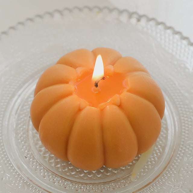 YIMWNYI Halloween Scented Candle, a soy wax aromatherapy candle shaped like a pumpkin with a vibrant orange color, burning on a clear textured glass plate. Compact at 7x7x5cm, it offers stress relief and relaxation, ideal for Halloween decor, fall home decor, or as a unique gift for women.