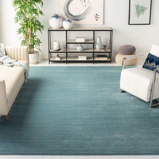 Safavieh Vision Adrasteia Overdyed Solid Area Rug in blue with subtle horizontal texture, placed in a contemporary living room featuring a cream sofa, white armchair with navy pillow, black metal shelving with decorative vases, a tall green plant, and abstract wall art. Suitable for modern urban or family room settings.