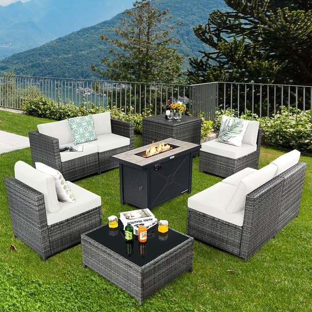 Gymax 9 PCS Patio Rattan Furniture Set with a black fire pit table offering 60,000 BTU heat output, off-white cushions, and durable PE rattan. Includes armless and corner sofas, a coffee table, and a storage box, ideal as an outdoor patio conversation set. Set in a lush garden with scenic mountain views.