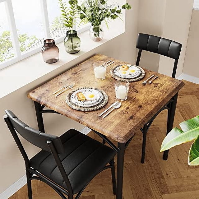 Dining Table Set for 2 featuring a square engineered wood table with a brown wood grain finish and two black upholstered chairs. The set is designed for small spaces like apartments or studios, with dimensions of 29.28 x 29.28 x 30.08 inches for the table. The chairs include soft black cushions for comfort and can be stowed under the table to save space. The metal frame and MDF boards ensure stability and durability, supporting up to 150 pounds for the table and 300 pounds for each chair. The set is shown in a bright dining area with natural light and decorative accents.