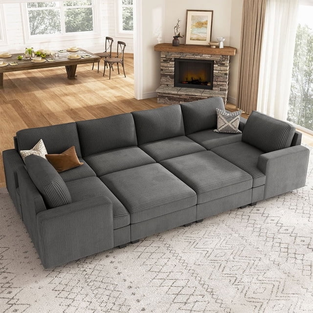 HONBAY Canapé Sectionnel-Lit Unisexe en Velours Côtelé Gris avec Rangement Réversible pour Salon. Ce Sofa Modulaire Présente un Tissu en Velours Côtelé Gris Foncé, Offrant une texture Douce et Agréable pour la Peau. Il Comprend Deux ottomans de Rangement Spacieux et une Housse Amovible et Lavable pour un Entretien Facile. le design Sectionnel Permet une Personnalisation et Se Transforme en Lit, Idéal pour Accueillir des Invités Ou Se Détendre. Montré dans un Salon Douillet avec un Foyer et un Coin Repas en Arrière-Plan.