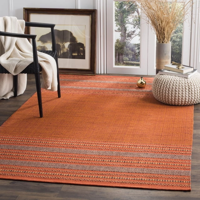 Safavieh Montauk Jobeth Striped Soft Braided Area Rug in flat-weave cotton with orange and beige stripes, shown on dark hardwood flooring in a bright room featuring a black chair with a cream knit throw, framed elephant art, and a knitted pouf with books and a gold decorative piece, suitable for coastal and contemporary décor.