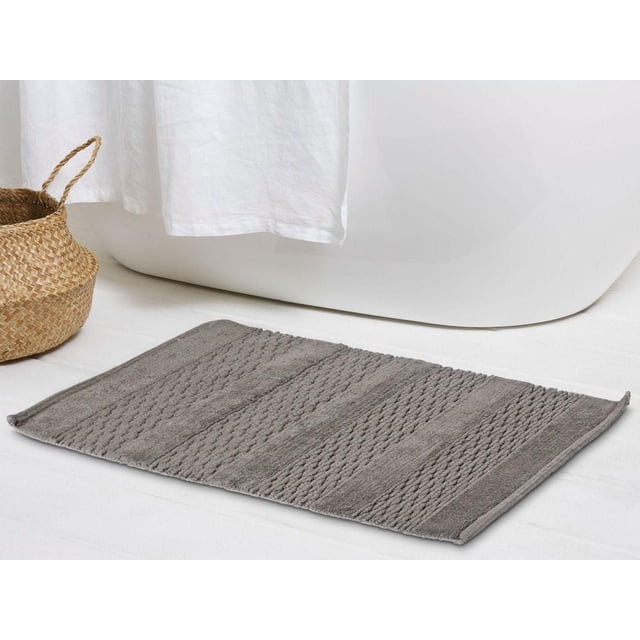 Regal Bath Rug Dark Grey 20 x 30 inches rectangular 100% cotton bath rug with a pattern of solid and woven textured stripes, placed on a white bathroom floor near a white bathtub and woven basket, machine washable and suitable for bathroom use.
