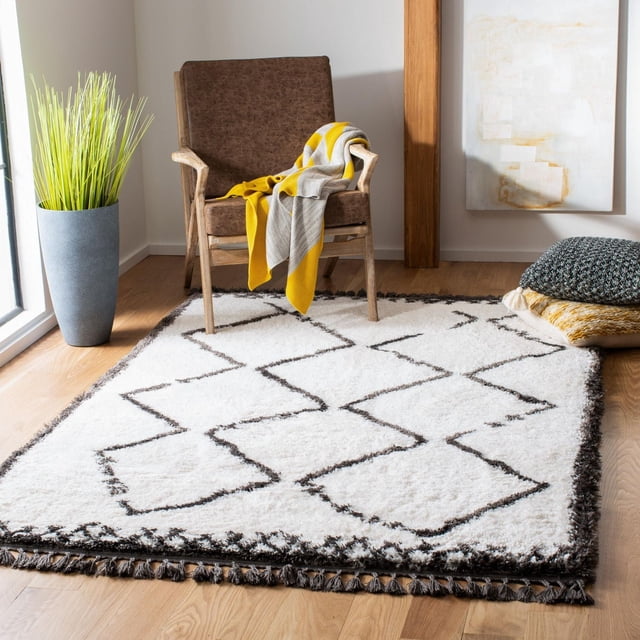 SAFAVIEH Carmella Stars Aztec Fringe Shag Area Rug featuring a plush white synthetic yarn pile with black geometric Aztec patterns and fringed edges, displayed on light hardwood flooring in a contemporary room with a brown wooden armchair draped with a yellow and gray throw, a tall gray planter with green grass, and stacked textured cushions, suitable for enhancing comfort and style in accent rug settings.