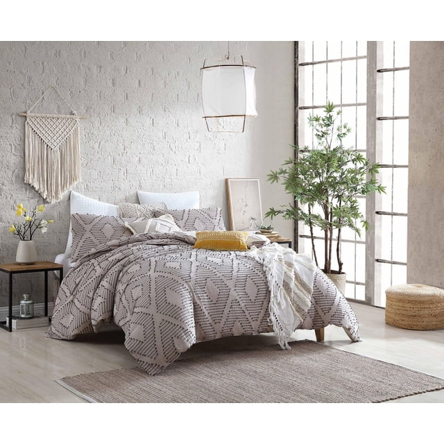 Cotton 5 Piece Puruma Comforter Set on a queen bed with beige cotton comforter featuring black geometric patterns, two matching pillow shams, a white square pillow, and an oblong decorative pillow. Displayed in a bright bedroom with white brick wall, hanging lantern, wooden side table with yellow flowers, large green plant by tall windows, and woven pouf on light hardwood floor with neutral rug. The set is machine washable, Oeko-Tex certified, and ideal for adding comfort and style to guest rooms.