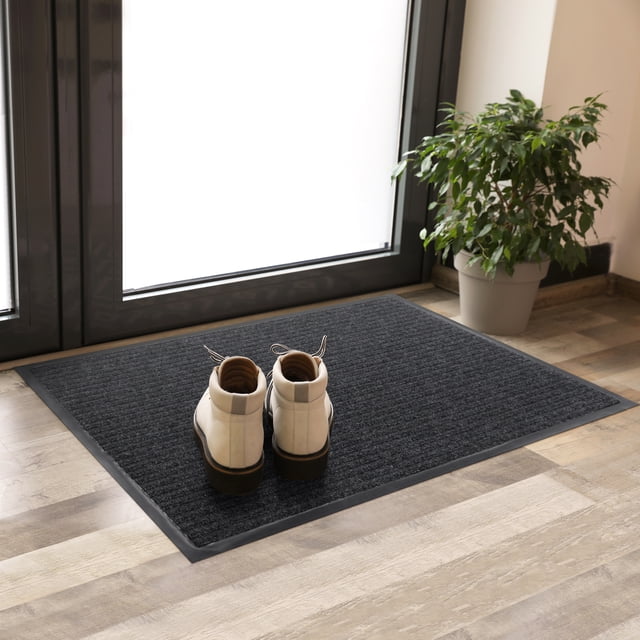 Stepguard 2'X3' Charcoal Needlepunch Ribbed Mat on a light wood floor by a glass door, featuring a ribbed recycled polyester surface with a phthalate-free vinyl border, holding a pair of white boots, ideal for indoor floor protection against dirt and moisture, also suitable for entryway use.