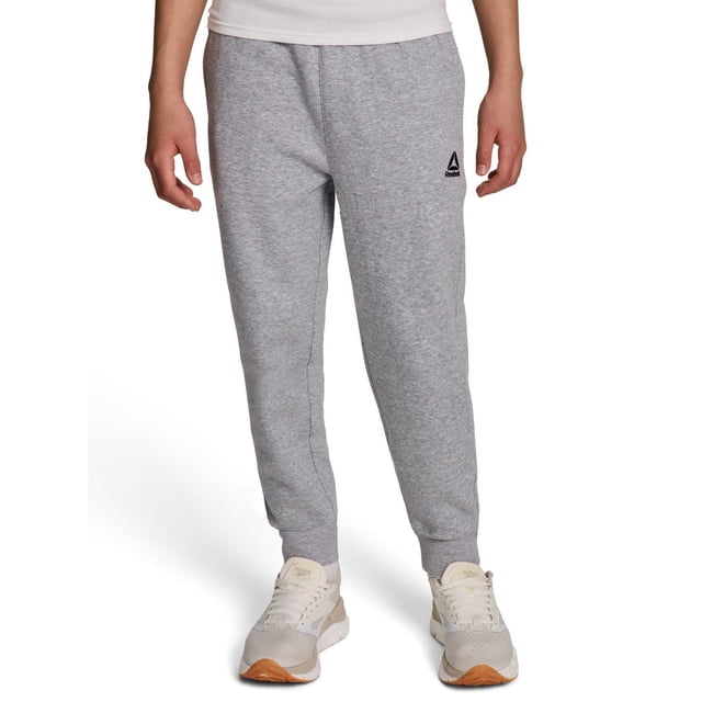 Reebok Boys Delta Fleece Jogger in light grey heather made of cotton-polyester fleece, with elastic waistband and inner drawcord, ribbed cuffs, two side pockets, and black Reebok Delta logo on left thigh, worn by a boy with white and beige sneakers, ideal for boys' activewear bottoms and casual wear.