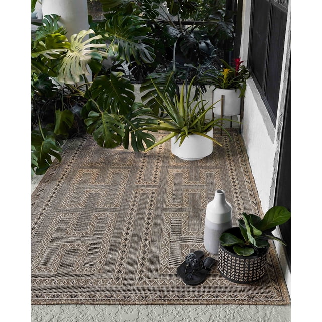 ECARPET Hazel Tapis Transitionnel pour Aire avec un Motif Géométrique Tissé Beige et Crème Neutre sur un Plancher Extérieur Texturé, Entouré de Plantes en Pot Vertes Incluant monstera et aloe, Accentué d'Un vase Décoratif Blanc et Gris et de Sandales Noires, Idéal pour les Espaces Intérieurs Ou Extérieurs.