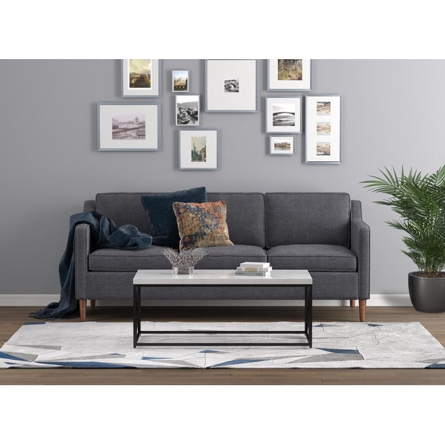 Safdie & Co. Table Basse 44 L Marbre Noir Métal avec Dessus Rectangulaire en Marbre Blanc et Cadre en Métal Noir, Placée devant un sofa Trois Places Gris Foncé avec Pattes en Bois et Coussins Décoratifs. la Table Repose sur un Tapis à Motif Géométrique dans un Salon avec un Mur Gris Pâle Présentant des Oeuvres Encadrées et une Plante Verte en Pot, Complétant le Mobilier de Salon comme Table d'Appoint Moderne.