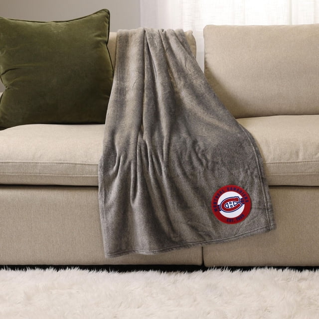 NHL, NBA, MLB Grey Sports Throw Blanket, 50" x 60", draped over a beige sofa with a green cushion, made of soft plush polyester with a Montreal Canadiens logo patch in red and blue, ideal for cozy indoor use and as a warm layering blanket.