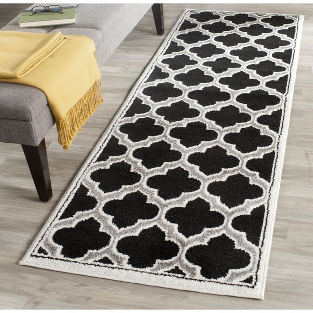 Safavieh Amherst Wilson Geometric Area Rug with black and white trellis pattern, power loomed polypropylene construction for durability in high-traffic indoor-outdoor spaces, displayed on light wood flooring next to a gray bench with a yellow throw and book with glasses, often used to enhance entryways or hallways.