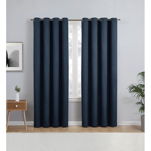 Eclipse Heath 63" grommet blackout window curtains panel in indigo blue, made of 100% polyester with light-blocking fabric, hanging on a metal rod in a room with light grey walls, wooden floor, a wooden side table with a white vase and plant, and a potted green plant, ideal for privacy, noise reduction, and energy savings.