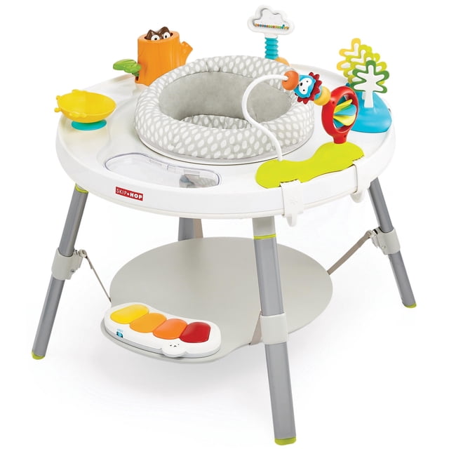 Skip Hop Explore n More 3 Stage Activity Center with a white circular frame and grey padded seat featuring white leaf patterns. It includes multicolour interactive toys such as a yellow spinning bowl, orange and red piano keys on a footrest, a blue tree, a cloud bead maze, and an orange tree stump with a brown owl figure. The activity center stands on four grey legs with lime green tips and has a green leaf-shaped play mat section for baby play and development.