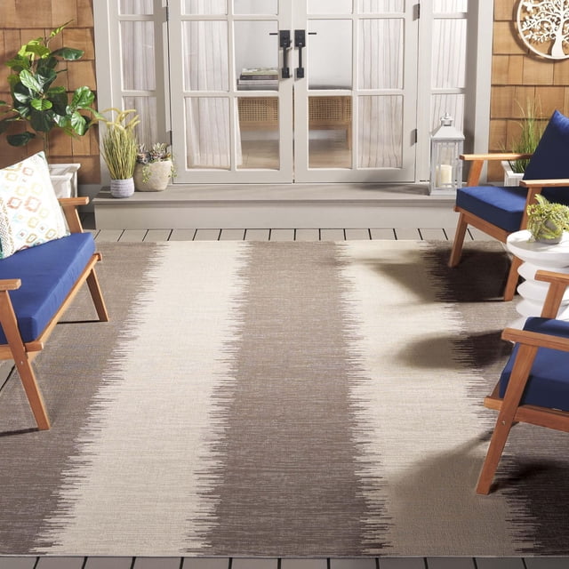 SAFAVIEH Courtyard Alexa Striped Indoor/Outdoor Area Rug with wide vertical beige, taupe, and brown stripes in a durable polypropylene sisal weave, placed on a wooden deck patio with blue cushioned wooden chairs, potted plants, and white French doors in the background, suitable for both indoor and outdoor use.