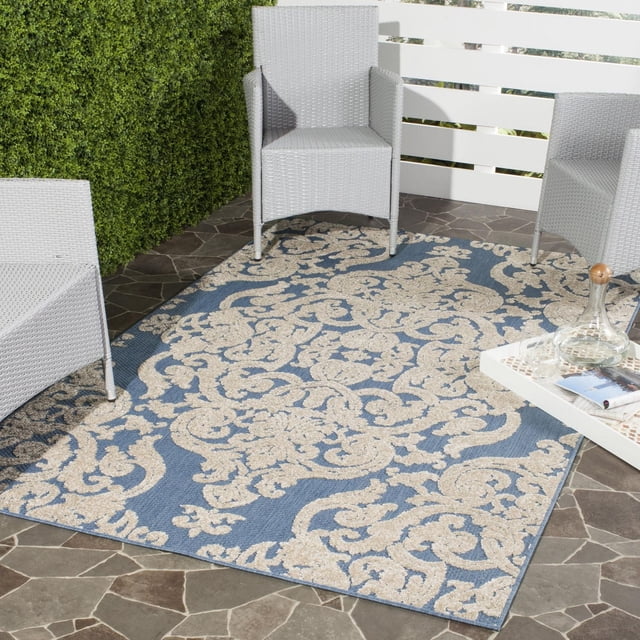 Safavieh Monroe Clarence Floral Area Rug in blue with beige floral scroll pattern, machine loomed from durable synthetic yarns, placed on stone patio with white wicker chairs and a white tray holding a glass decanter and glasses, ideal for indoor or outdoor use.