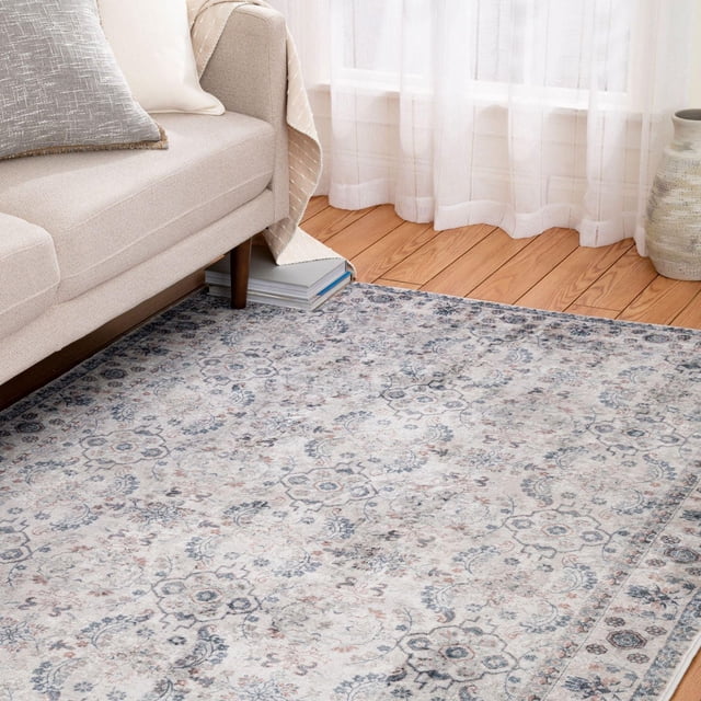 Banburry Light Grey and Dark Grey Woven Area Rug with a faded vintage floral pattern in grey tones, placed on a light hardwood floor beside a beige sofa with cushions and a throw, illuminated by sheer white curtains, ideal for living rooms or home offices.