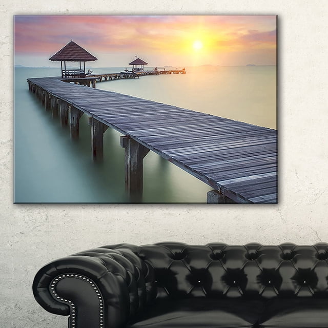 Design Art Wooden Sea Bridge And Sunset Seashore Photo Canvas Art Print showing a gallery-wrapped cotton canvas with a wooden pier extending over calm water toward two thatched roof gazebos under a soft orange and pink sunset sky, printed with high-resolution giclée inks and ready to hang with included hardware, suitable for adding a serene coastal touch to home or office walls.