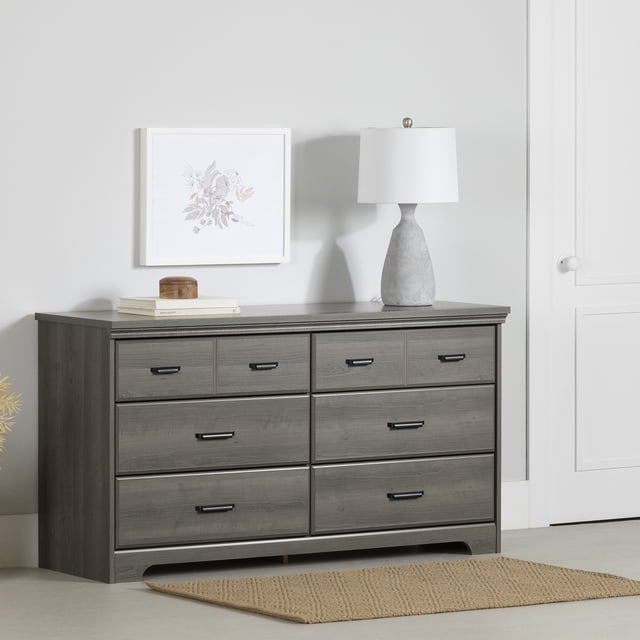 South Shore Versa 6-Drawer Dresser for Adult in gray maple finish with six spacious drawers featuring black metal handles, beveled edges, and a curved kick plate, positioned against a light gray wall with framed floral artwork, topped with a concrete-textured lamp and books, on a beige woven rug, ideal for organized storage in compact bedrooms.
