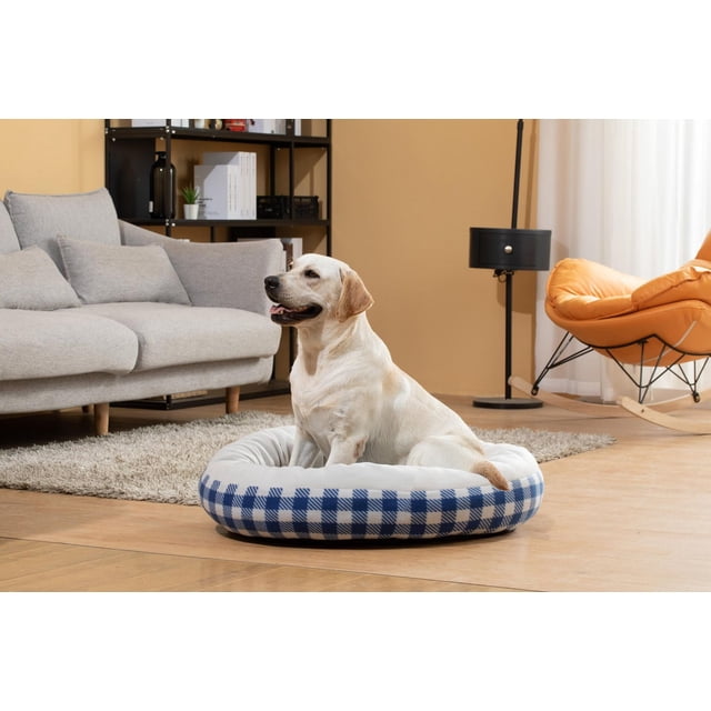 Pet Space Round Bed Red with soft polyester cushion and polypropylene bottom, shown in a blue and white checkered pattern with a Labrador dog sitting inside on a wooden floor in a living room with a grey sofa and orange rocking chair, suitable for providing a comfortable sleep area for dogs.