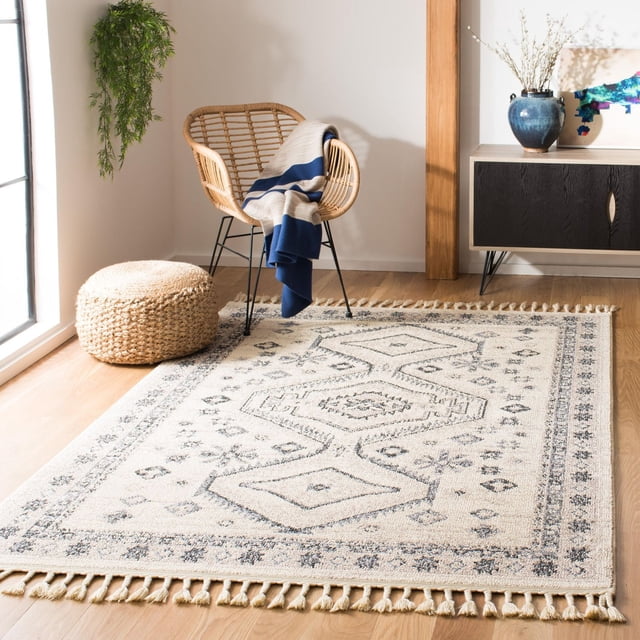 Safavieh Marrakesh Antilla Southwestern Fringe Area Rug with beige base and dark geometric tribal motifs, featuring fringed edges. Displayed on light hardwood flooring in a sunlit room with a rattan chair holding a blue and beige throw, woven pouf, and a black and wood sideboard with a blue vase. Ideal for adding boho-chic texture and warmth to living spaces.