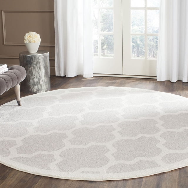 Safavieh Amherst Roderick Geometric Area Rug, a round indoor-outdoor accent rug with a light beige and cream geometric pattern, placed on dark hardwood flooring in a bright room with white curtains and a gray side table holding a white vase of roses.