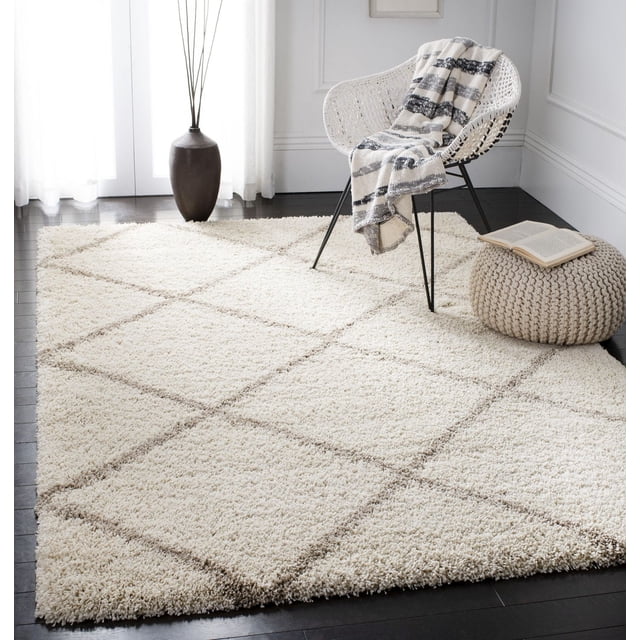 Safavieh Hudson Tapis Shag Géométrique Amias Mettant en Vedette une Épaisse pile Shag Ivoire avec de Subtiles Lignes de Motif de Carreaux Géométriques sur des Planchers de Bois Franc Sombres, Style avec une Chaise Tissée Blanche Drapée d'Un Jeté à Rayures, un pouf Tricoté beige Tenant un Livre Ouvert, et un Grand vase Foncé avec des branches Décoratives dans une Pièce Doucement Éclairée. Ce Tapis Peluche Ajoute une texture Réconfortante au décor Moderne et Convient aux Espaces de Vie Très Passants.