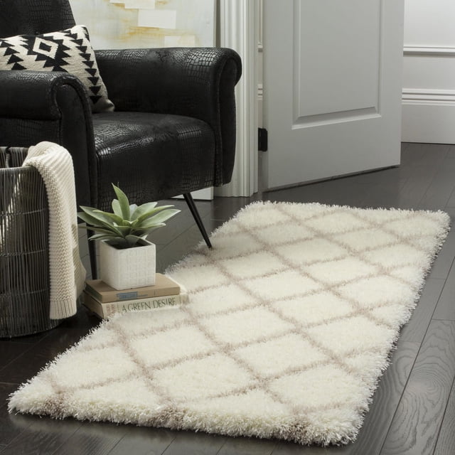 Safavieh Montreal Vesela Geometric Shag Area Rug in cream with beige diamond pattern, featuring plush pile texture on dark hardwood floor beside a black textured armchair with a geometric pillow, a potted plant on stacked books, and a woven basket with a cream throw, suitable as an accent rug for contemporary interiors.