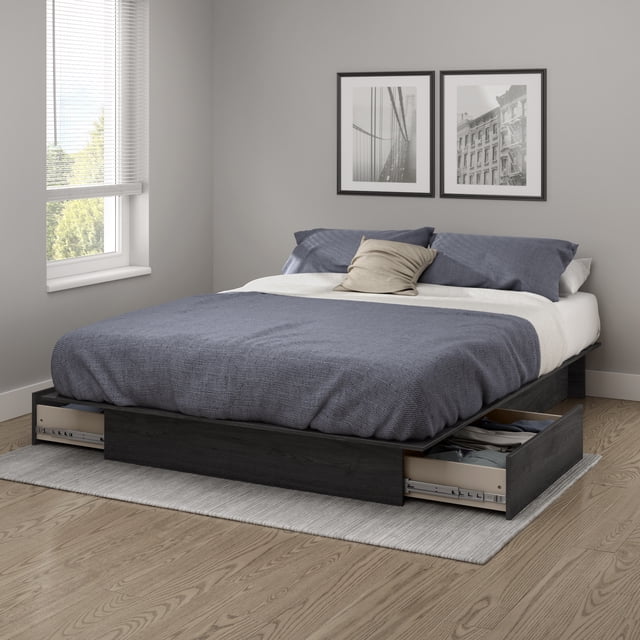 South Shore Step One Full/Queen Platform Bed with drawers in gray oak, shown in a modern bedroom with gray walls and wood flooring. The bed features a low-profile frame with two open underbed drawers on each side revealing folded clothes. It supports a mattress with a blue textured blanket, two blue pillows, and a beige accent pillow. The room includes a window with white blinds and two black-framed monochrome photos above the bed. The drawers have smooth ball-bearing full-extension slides for quiet, easy access, ideal for maximizing bedroom storage and organization.