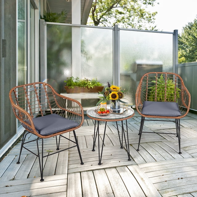 Costway 3PCS Patio Rattan Bistro Set with two armchairs featuring gray cushions and a round tempered glass coffee table. The compact and lightweight set is displayed on a wooden patio surrounded by greenery and sunflowers, suitable for gardens, balconies, and poolside areas.