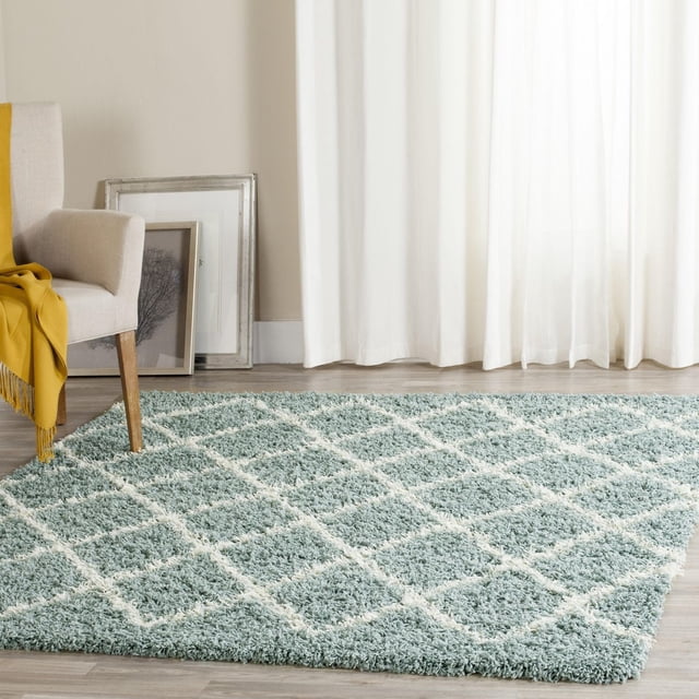 Safavieh Dallas Jerrie Geometric Shag Area Rug with a plush blue synthetic fiber pile and white diamond pattern, shown on light wood flooring beside a beige armchair with a mustard yellow throw, suitable for high-traffic home areas and adding texture to modern interiors.